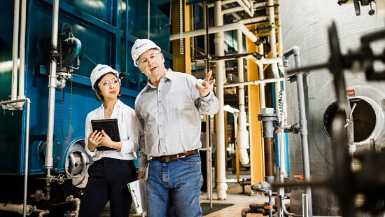 Two workers in safety gear inspect industrial pipes and tanks, one holding a tablet while the other gestures.